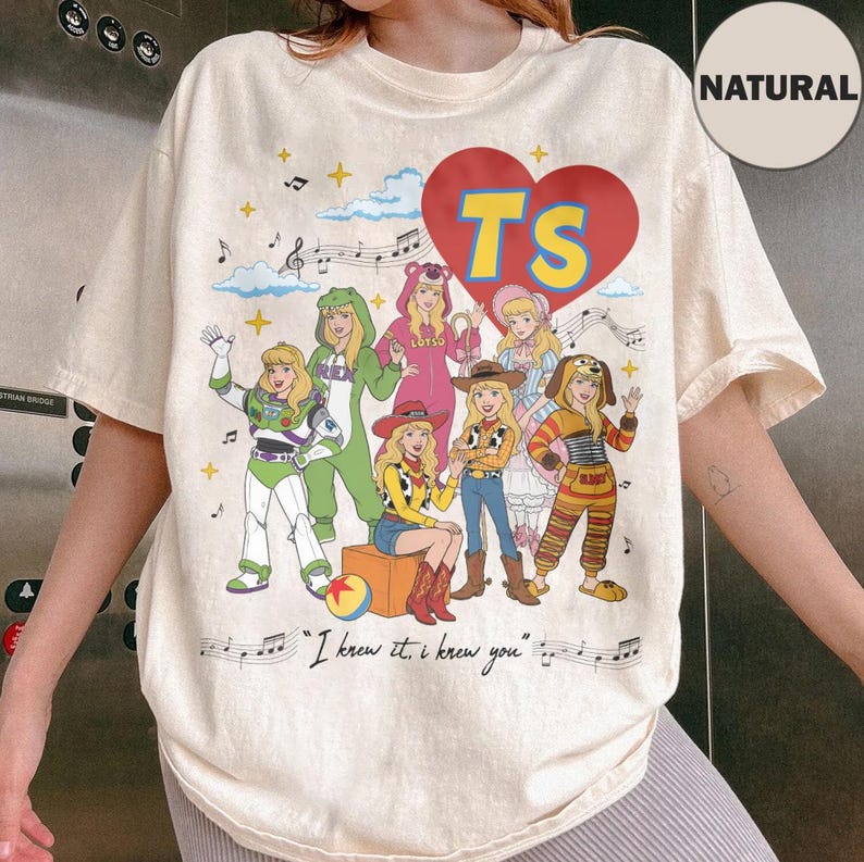 May include: A natural-colored t-shirt featuring a graphic of six characters dressed in various costumes, a large red heart with the yellow text TS, musical notes, and the text I knew it, I knew you printed in black script at the bottom.