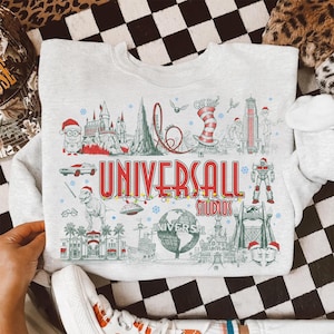 May include: A light gray sweatshirt featuring a colorful graphic of Universal Studios attractions and characters, including a roller coaster, a dinosaur, and the Cat in the Hat. The text "UNIVERSALL STUDIOS" is displayed in red.