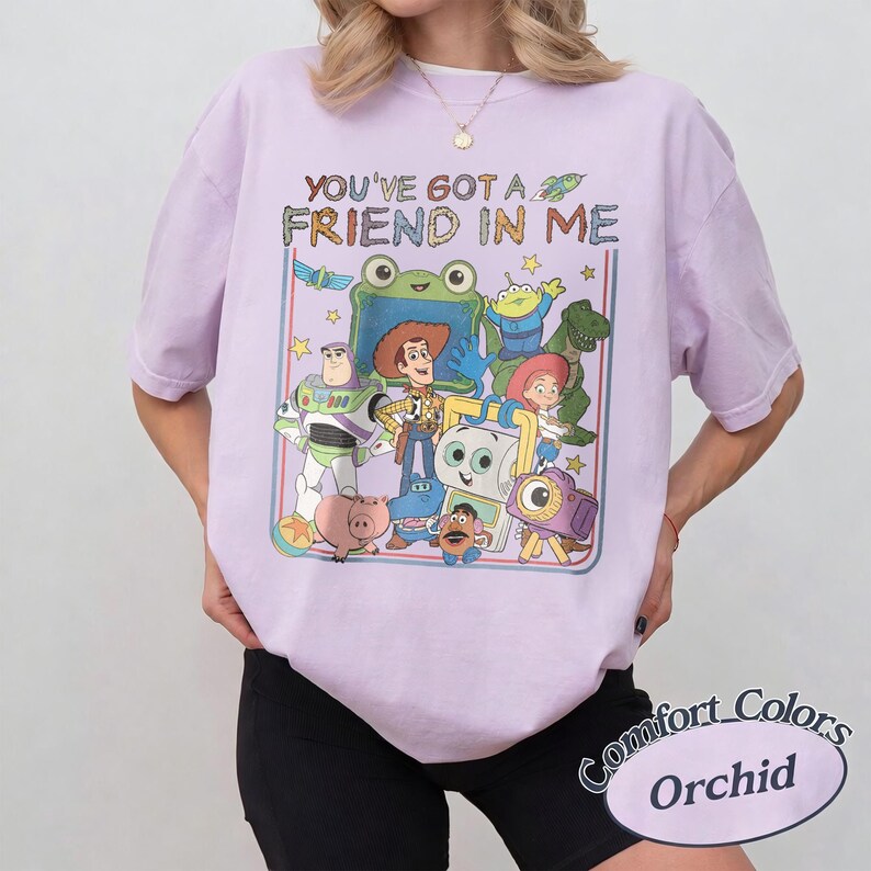 May include: A light purple short-sleeve t-shirt featuring a graphic print of various animated characters from the Toy Story franchise. The text "YOU'VE GOT A FRIEND IN ME" is printed above the characters in a multi-colored, textured font style.