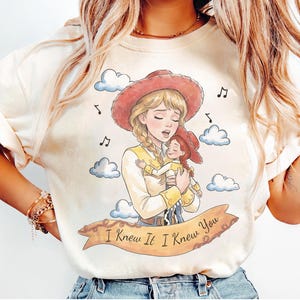 May include: A cream-coloured t-shirt featuring a graphic illustration of a blonde cowgirl holding a smaller cowgirl doll. The design includes blue clouds, musical notes, and a tan banner with the text "I Knew It I Knew You" in black script.