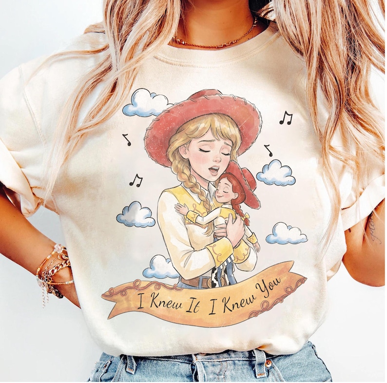 May include: A cream-coloured t-shirt featuring a graphic illustration of a blonde cowgirl holding a smaller cowgirl doll. The design includes blue clouds, musical notes, and a tan banner with the text "I Knew It I Knew You" in black script.