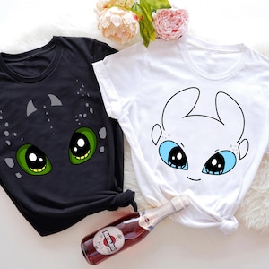 May include: Two t-shirts, one black and one white, each with a cartoon dragon face. The black shirt has green eyes and grey accents. The white shirt has blue eyes and black outlines. Both are short-sleeved and knotted at the bottom.