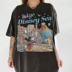 Pu&ograve; includere: T-shirt nero con una grafica colorata di Topolino in kimono, un castello e la scritta "Tokyo Disney Sea". Il design include un globo, un vulcano e dei palloncini. La maglietta ha un aspetto vintage e usurato.