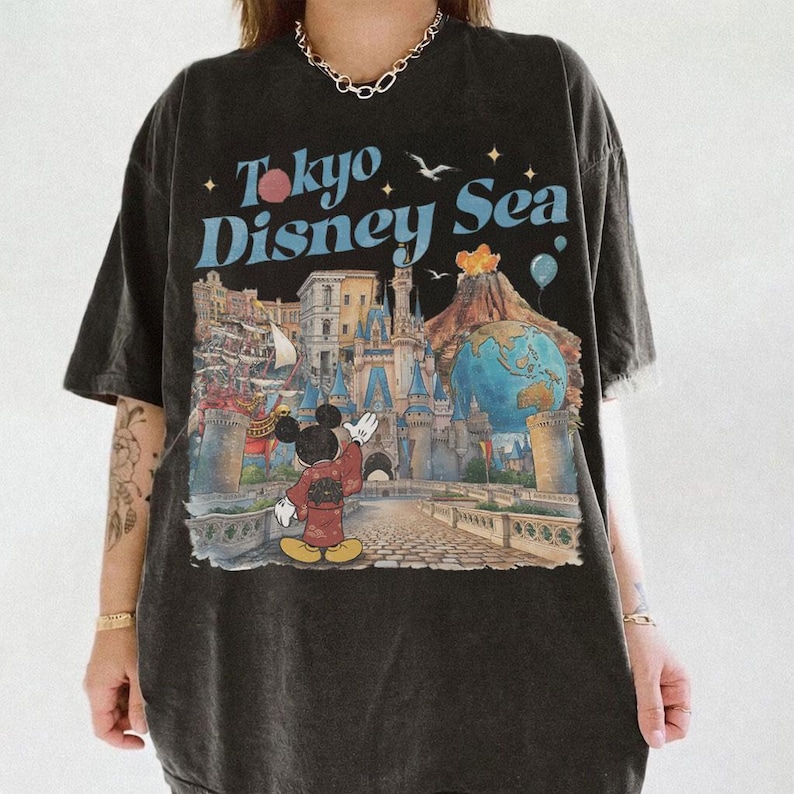 Pu&ograve; includere: T-shirt nero con una grafica colorata di Topolino in kimono, un castello e la scritta "Tokyo Disney Sea". Il design include un globo, un vulcano e dei palloncini. La maglietta ha un aspetto vintage e usurato.