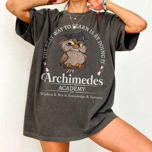 May include: A charcoal gray t-shirt featuring a graphic of an owl wearing a graduation cap. The text reads: THE BEST WAY TO LEARN IS BY DOING IT, Archimedes ACADEMY OF Wisdom & Wit & Knowledge & Sarcasm, with two small diploma scrolls.