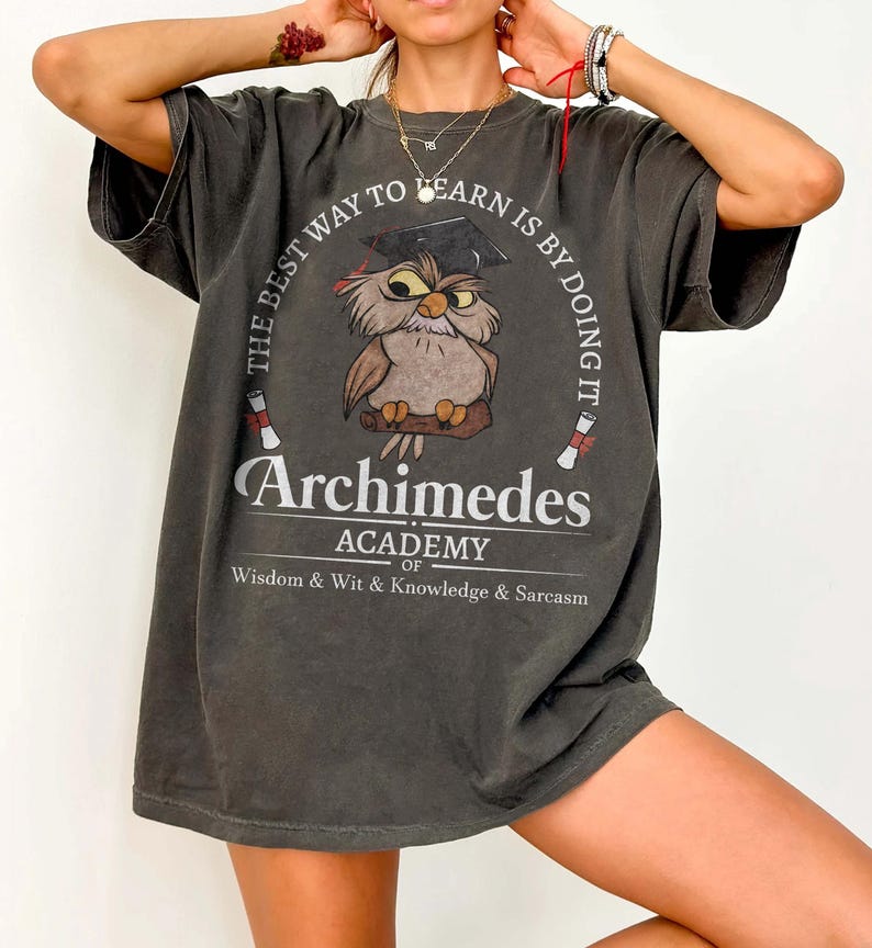 May include: A charcoal gray t-shirt featuring a graphic of an owl wearing a graduation cap. The text reads: THE BEST WAY TO LEARN IS BY DOING IT, Archimedes ACADEMY OF Wisdom & Wit & Knowledge & Sarcasm, with two small diploma scrolls.