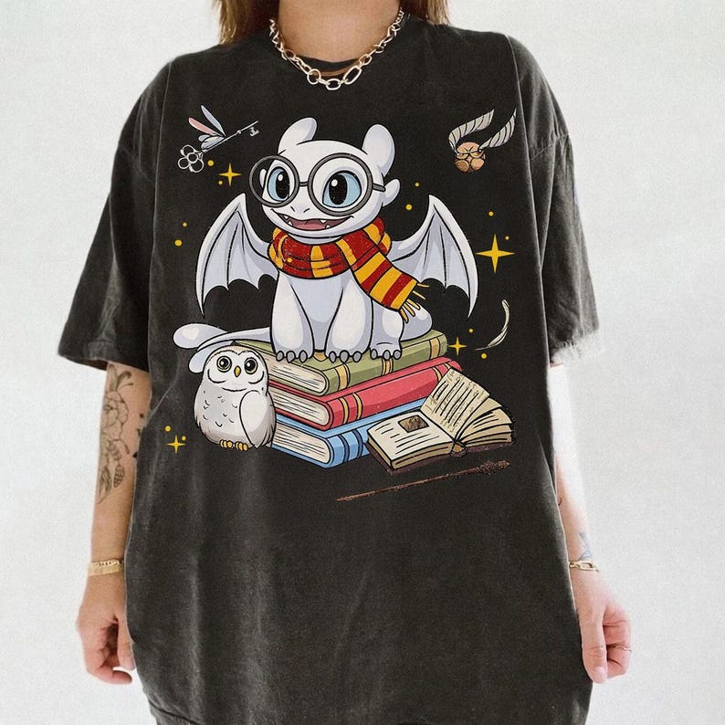 May include: Dark gray t-shirt featuring a cartoon dragon wearing glasses and a scarf, sitting on books. An owl and other whimsical elements are also depicted. The design is in a cartoon style with bright colors and star accents.