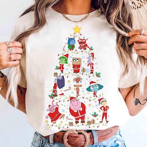 May include: A cream-colored t-shirt featuring a Christmas tree design made of cartoon characters. The tree is decorated with lights, stars, and gifts. The characters are in festive outfits, with a yellow star at the top.