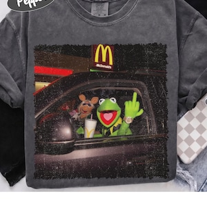 May include: A charcoal gray crew neck t-shirt featuring a graphic print of Kermit the Frog and Miss Piggy in a car at a McDonald's drive-thru. Kermit is shown with a heart-shaped mouth, wearing a gold watch, and gesturing with a middle finger.