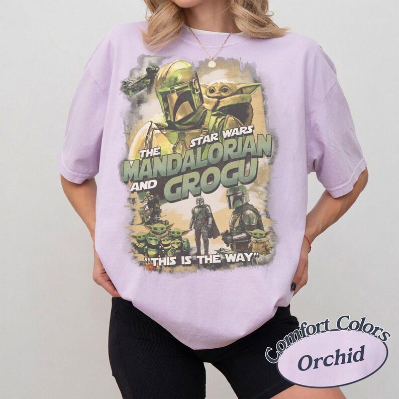 Może przedstawiać: Jasnofioletowa koszulka z grafiką Star Wars z The Mandalorian i Grogu. Projekt zawiera obrazy The Mandalorian, Grogu i innych postaci z napisem "THIS IS THE WAY". Koszulka to Comfort Colors Orchid.