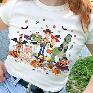 Puede incluir: Camiseta de color crema con un diseño de Halloween de Toy Story. El diseño presenta a Woody, Buzz Lightyear, Jessie, Rex y otros personajes, junto con calabazas, murciélagos y hojas de otoño.