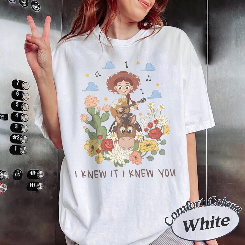 Op de afbeelding: Een wit t-shirt met een afbeelding van een cartoon cowgirl die gitaar speelt op een paard, omringd door cactussen, bloemen, wolken en muzieknoten. De tekst "I KNEW IT I KNEW YOU" is in het bruin onder de illustratie gedrukt.