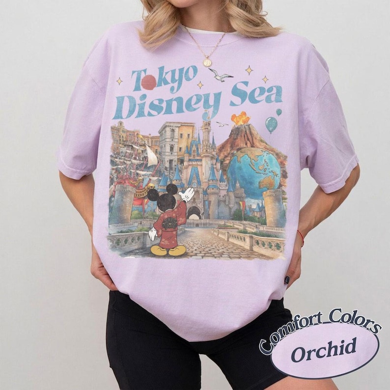 Pu&ograve; includere: Una maglietta lilla con la scritta "Tokyo Disney Sea" in blu e una grafica di Topolino davanti a un castello e un vulcano. La maglietta &egrave; etichettata "Comfort Colors Orchid".