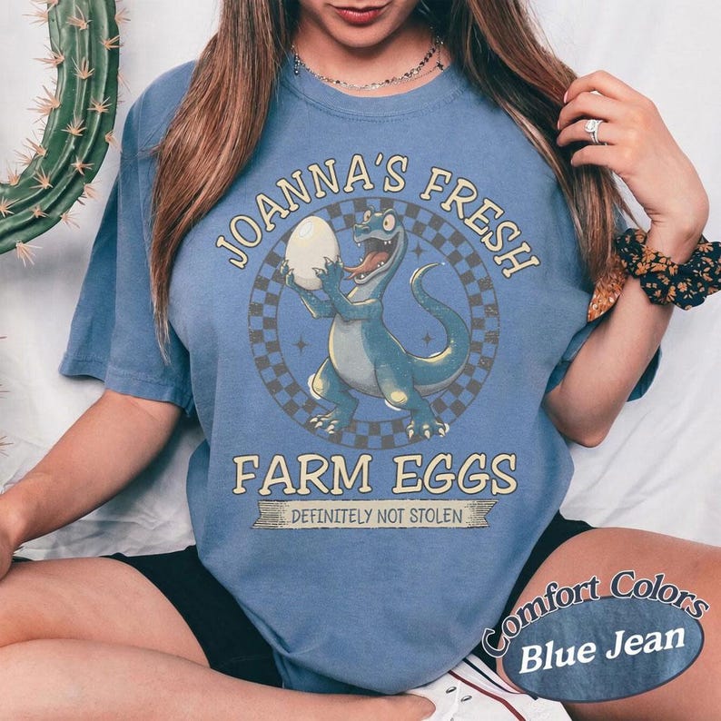 May include: Blue jean colored t-shirt with a graphic of a cartoon dinosaur holding an egg. Text on the shirt reads "JOANNA'S FRESH FARM EGGS" and "DEFINITELY NOT STOLEN". The shirt is a Comfort Colors brand.