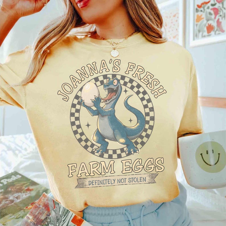 May include: Yellow t-shirt with a dinosaur graphic holding an egg, surrounded by text that reads "JOANNA'S FRESH FARM EGGS - DEFINITELY NOT STOLEN." The dinosaur is blue, and the egg is white. The shirt is a light yellow color.
