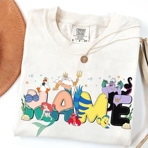 Puede incluir: Camiseta color crema con la palabra "NAME" en letras coloridas de estilo caricatura con personajes de Disney. La camiseta incluye a Ariel, Flounder, Sebastián y Úrsula. También se ven un collar dorado y un sombrero marrón.
