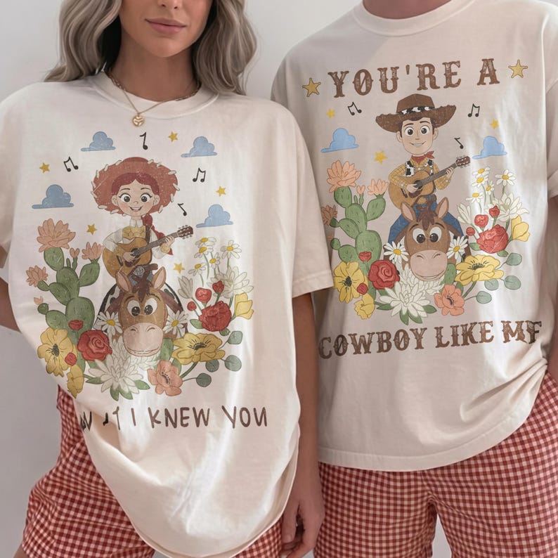 Op de afbeelding: Twee cr&egrave;mekleurige t-shirts met grafische prints van stripfiguren die gitaar spelen. Het linker t-shirt toont een cowgirl en paard met de tekst "I KNEW YOU". Het rechter t-shirt toont een cowboy en paard met "YOU'RE A COWBOY LIKE ME".