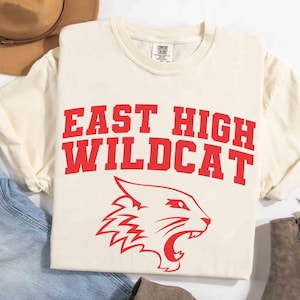 Op de afbeelding: Crèmekleurig T-shirt met de tekst "EAST HIGH WILDCAT" in rode blokletters en een rode wilde kat-afbeelding. Het shirt wordt getoond met een hoed, jeans en laarzen.