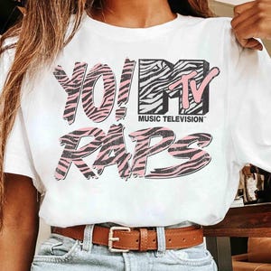 May include: White t-shirt featuring the text "YO! MTV RAPS" in a pink and zebra print design. The MTV logo is also present in black and pink. The shirt is paired with blue jeans and a brown belt.