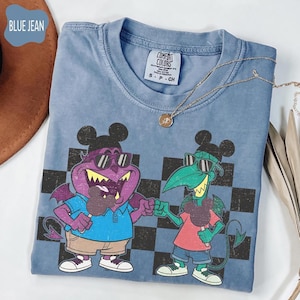 May include: Blue jean colored t-shirt featuring a cartoon graphic of two characters with Mickey Mouse ears, sunglasses, and holding ice cream bars. The shirt has a crew neck and the brand name "Comfort Colors" is visible. A gold necklace is draped across the shirt.