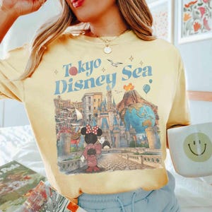 Pu&ograve; includere: T-shirt giallo chiaro con la scritta "Tokyo Disney Sea" e un disegno di Minnie Mouse in kimono, un castello, un vulcano e un globo. Il design &egrave; in stile vintage.