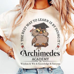 May include: A cream-colored t-shirt featuring a graphic of a brown owl wearing a black graduation cap. The text reads: THE BEST WAY TO LEARN IS BY DOING IT, Archimedes ACADEMY OF Wisdom & Wit & Knowledge & Sarcasm, with two small scrolls.
