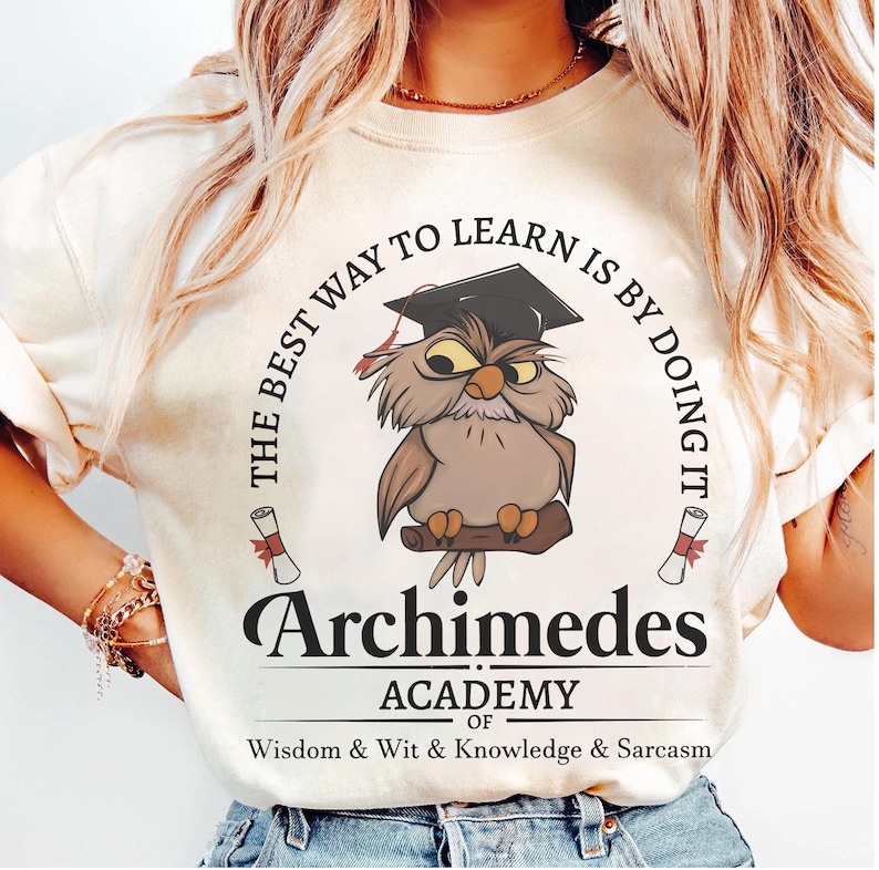 May include: A cream-colored t-shirt featuring a graphic of a brown owl wearing a black graduation cap. The text reads: THE BEST WAY TO LEARN IS BY DOING IT, Archimedes ACADEMY OF Wisdom & Wit & Knowledge & Sarcasm, with two small scrolls.