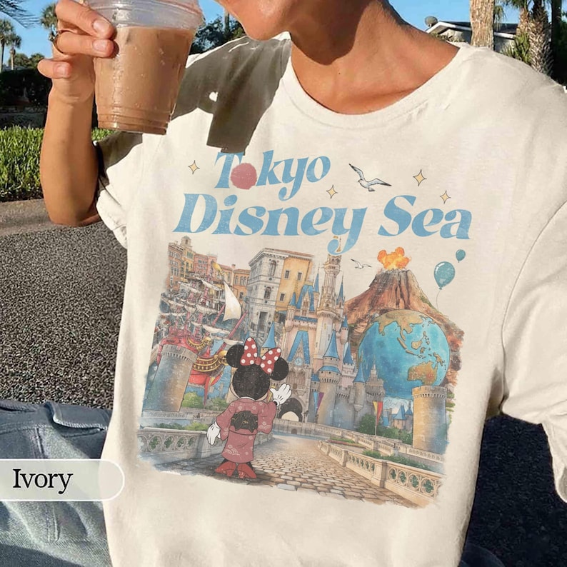 Pu&ograve; includere: T-shirt color avorio con la scritta "Tokyo Disney Sea" in blu e rosa. La grafica include Minnie Mouse in kimono, un castello, un vulcano e un globo. Un capo d'abbigliamento casual.