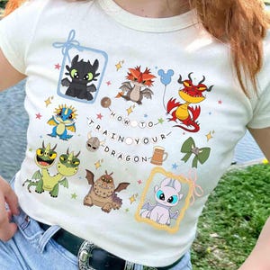 May include: Off-white t-shirt featuring colorful cartoon dragons and the text "HOW TO TRAIN YOUR DRAGON." The shirt has various dragon illustrations, stars, and decorative elements. The shirt is being worn by a person.
