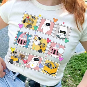 Può includere: T-shirt color crema con una griglia di immagini dei personaggi dei cartoni animati WALL-E ed EVE. Ogni immagine è incorniciata in un colore diverso, con palloncini a forma di Topolino e stelle. La maglietta è uno stile casual e divertente.