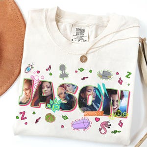 May include: Cream-colored t-shirt with the name "JASON" in large, colorful letters. The letters feature images of people and are surrounded by decorative elements like stars, musical notes, and a peace sign. The word "ZOMBIES" is below the name.