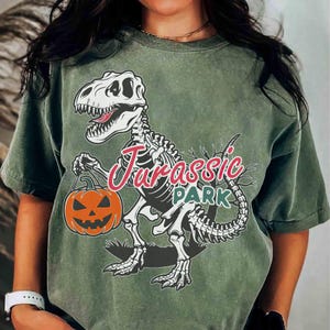 Peut inclure: T-shirt vert olive avec un dinosaure squelette tenant une citrouille orange. Le texte "Jurassic Park" est écrit en rose et vert, créant un design sur le thème d'Halloween.