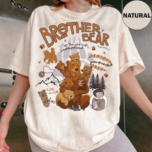 Pode incluir: Camiseta creme com um gráfico castanho com as palavras "Brother Bear" e ilustrações de ursos, montanhas e árvores. O texto "The Bonds of Brotherhood" também está incluído. A camisa tem mangas curtas e um ajuste folgado.