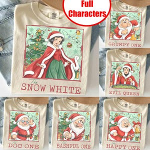 Könnte beinhalten: Ein cremefarbenes T-Shirt mit einer Grafik von Schneewittchen in einem rot-weißen Umhang im Weihnachtsmann-Stil vor einem Weihnachtsbaum. Der Text darunter lautet The SNOW WHITE. Weitere T-Shirts zeigen Figuren mit dem Text The GRUMPY ONE, The EVIL QUEEN, The DOC ONE, The BASHFUL ONE und The HAPPY ONE.