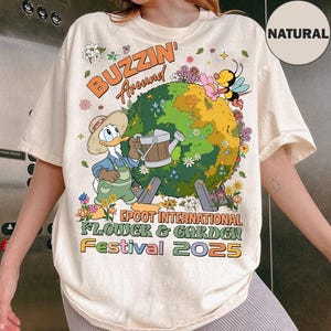 Puede incluir: Camiseta color crema con un diseño gráfico colorido. El diseño incluye el texto "BUZZIN' Around" sobre un globo con flores y un pato de dibujos animados regándolo. El texto "EPCOT INTERNATIONAL FLOWER & GARDEN FESTIVAL 2025" está debajo.