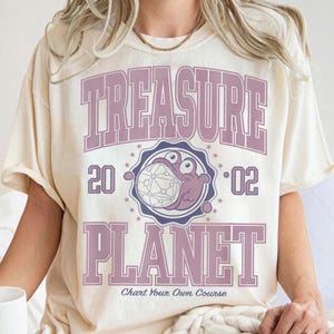 Könnte beinhalten: Ein cremefarbenes Kurzarm-T-Shirt mit dem Text TREASURE PLANET in großen violetten Blockbuchstaben. Die Grafik enthält die Zahlen 20 und 02, ein kreisförmiges Emblem mit einer Comic-Figur, die einen Globus hält, und den Text Chart Your Own Course.