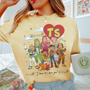 May include: A pale yellow t-shirt featuring a graphic of seven characters dressed in various costumes, a large red heart with the letters TS, musical notes, and the text "I knew it. I knew you." The design includes stars, clouds, and a colorful ball.