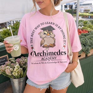 May include: A light pink t-shirt featuring a graphic of an owl wearing a graduation cap, with the text "THE BEST WAY TO LEARN IS BY DOING IT", "Archimedes", "ACADEMY OF", and "Wisdom & Wit & Knowledge & Sarcasm" printed in black ink.