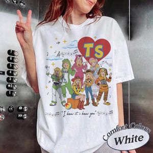 May include: A white t-shirt featuring a large red heart with the letters TS in yellow. Below the heart are seven illustrated characters dressed in various costumes, including a space ranger and a cowgirl, with musical notes and the text I knew it. I knew you.
