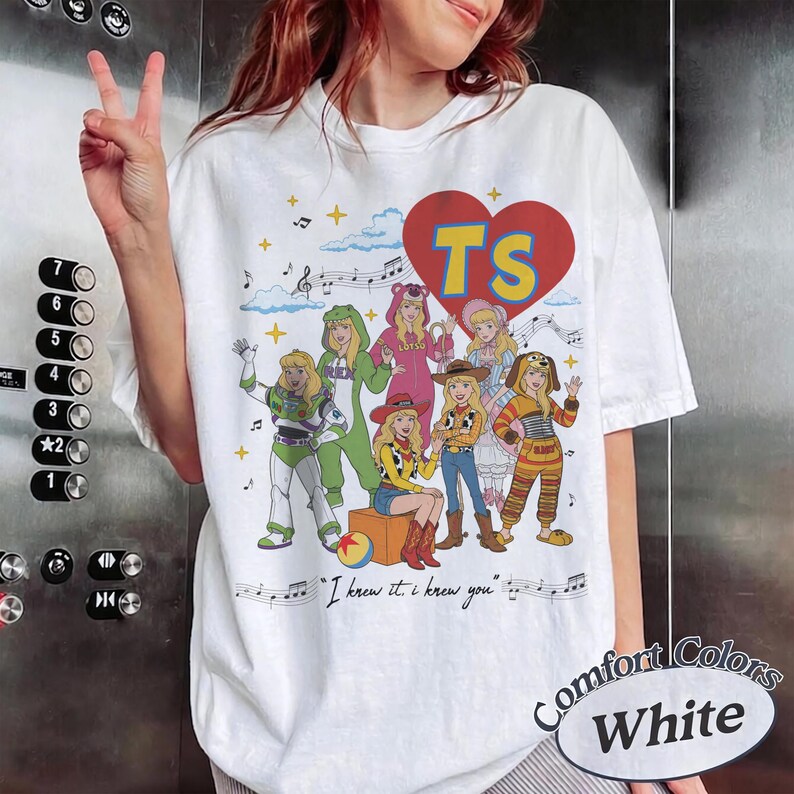 May include: A white t-shirt featuring a large red heart with the letters TS in yellow. Below the heart are seven illustrated characters dressed in various costumes, including a space ranger and a cowgirl, with musical notes and the text I knew it. I knew you.