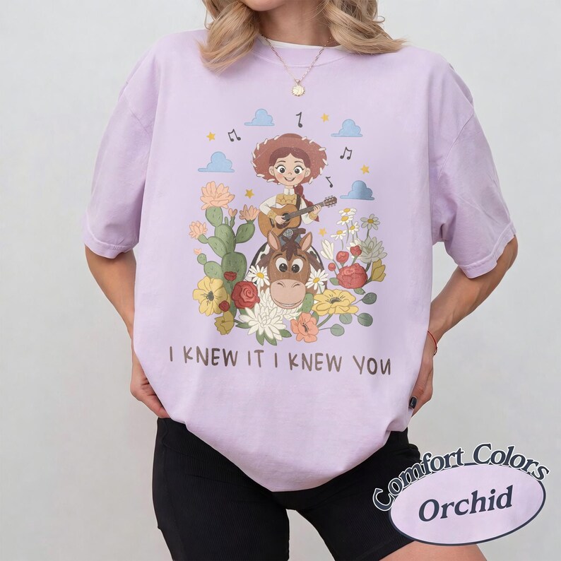 Op de afbeelding: Een lichtpaars t-shirt met een afbeelding van een cowgirl die gitaar speelt op een paard, omringd door cactussen en bloemen. De tekst "I KNEW IT I KNEW YOU" staat onder de illustratie gedrukt. Het t-shirt heeft korte mouwen.