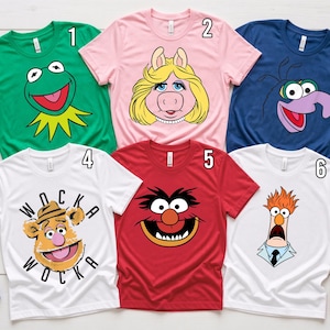 May include: Six short-sleeved t-shirts featuring Muppets characters: a green Kermit, pink Miss Piggy, blue Gonzo, white Fozzie Bear with the text WOCKA WOCKA, red Animal, and white Beaker. Each shirt is numbered one through six in the corner.