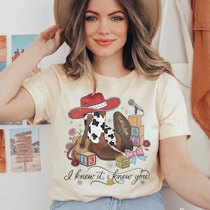 Pu&ograve; includere: Una t-shirt color crema con una stampa grafica raffigurante un cappello da cowboy rosso, stivali con stampa mucca, una chitarra acustica, un microfono, blocchi dell'alfabeto e fiori. Il testo "TAYLOR" &egrave; su uno stivale e "I knew it, i knew you." &egrave; sotto.