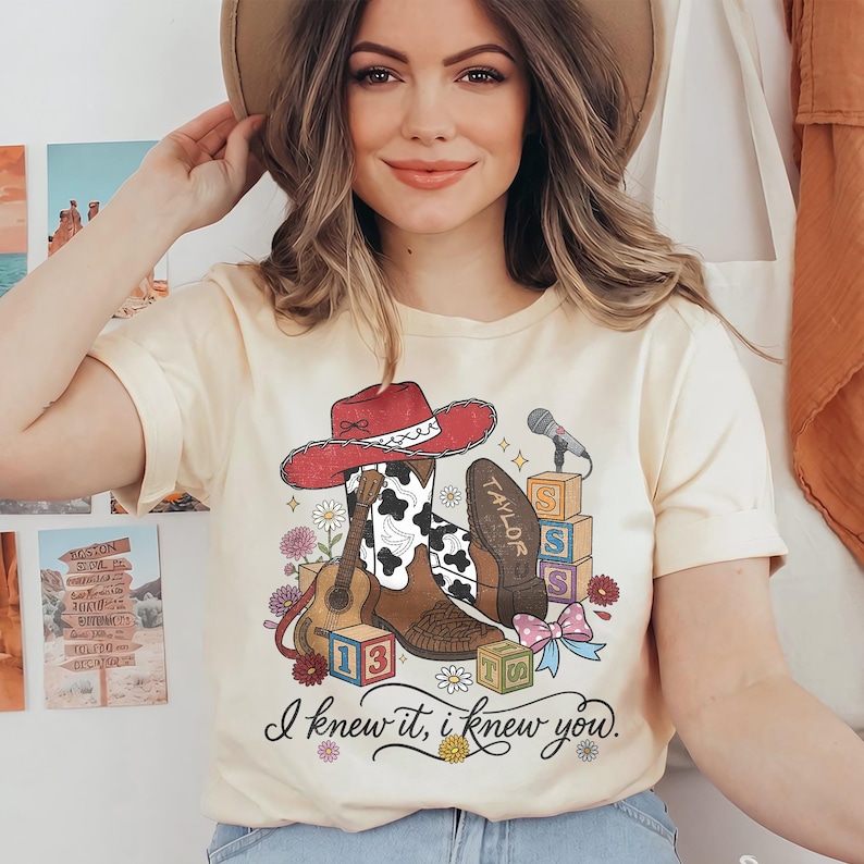 Pu&ograve; includere: Una t-shirt color crema con una stampa grafica raffigurante un cappello da cowboy rosso, stivali con stampa mucca, una chitarra acustica, un microfono, blocchi dell'alfabeto e fiori. Il testo "TAYLOR" &egrave; su uno stivale e "I knew it, i knew you." &egrave; sotto.
