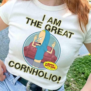 May include: Off-white t-shirt featuring a cartoon image of Beavis from Beavis and Butt-Head, with the text "I AM THE GREAT CORNHOLIO!". The graphic is in a circle with a red couch in the background. The shirt has a crew neck and short sleeves.