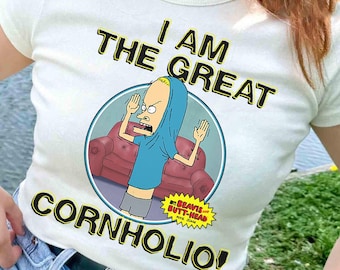 May include: Off-white t-shirt featuring a cartoon image of Beavis from Beavis and Butt-Head, with the text "I AM THE GREAT CORNHOLIO!". The graphic is in a circle with a red couch in the background. The shirt has a crew neck and short sleeves.