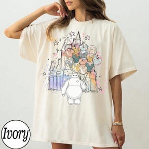 Puede incluir: Camiseta color marfil con una ilustraci&oacute;n colorida de Baymax sosteniendo globos de Mickey Mouse frente a un castillo. El dise&ntilde;o incluye estrellas y detalles de estilo acuarela. La camiseta tiene un corte holgado.