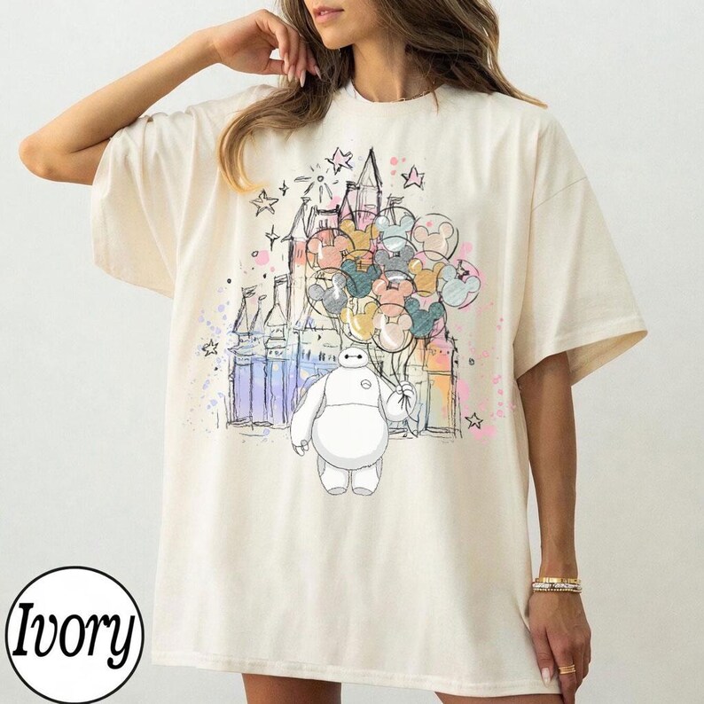 Puede incluir: Camiseta color marfil con una ilustraci&oacute;n colorida de Baymax sosteniendo globos de Mickey Mouse frente a un castillo. El dise&ntilde;o incluye estrellas y detalles de estilo acuarela. La camiseta tiene un corte holgado.