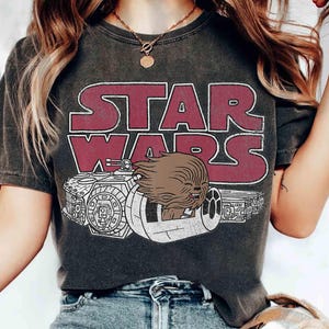 Puede incluir: Camiseta gris oscuro con el logo de Star Wars en burdeos y blanco. El gráfico incluye una imagen de Chewbacca en un vehículo. La camiseta es de estilo vintage y tiene un aspecto desgastado.