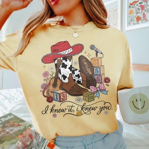 Pu&ograve; includere: Una t-shirt girocollo giallo pallido con una stampa grafica raffigurante un cappello da cowboy rosso, stivali con stampa mucca, una chitarra acustica, un microfono, cubi dell'alfabeto e un fiocco. Il testo recita "TAYLOR" e "I knew it, i knew you."
